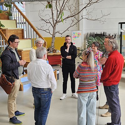 Blick auf eine im Kreis stehende Gruppe, die gerade eine Führung zur Präsenzstelle und dessen Makerspace mit Manuel Haberland bekommt.