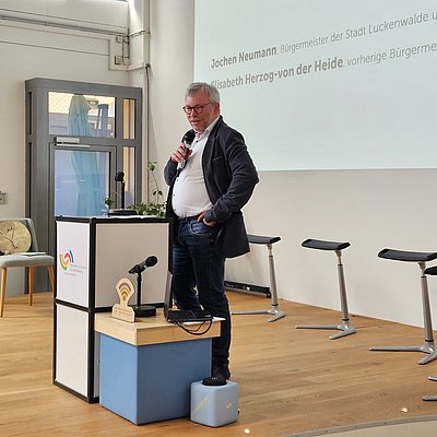Jochen Neumann, Bürgermeister der Stadt Luckenwalde, steht auf der Bühne und hält den zweiten Teil der Keynote zur aktuellen Wirkkraft der Präsenzstelle und die zukünftigen Pläne rund um die Präsenzstelle.